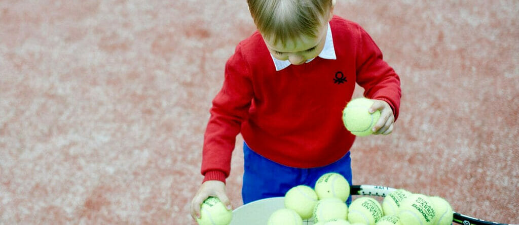 Kid on a Red Artificial Grass Tennis Court Collects Tennis Balls into the Bucket