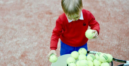 Kid on a Red Artificial Grass Tennis Court Collects Tennis Balls into the Bucket